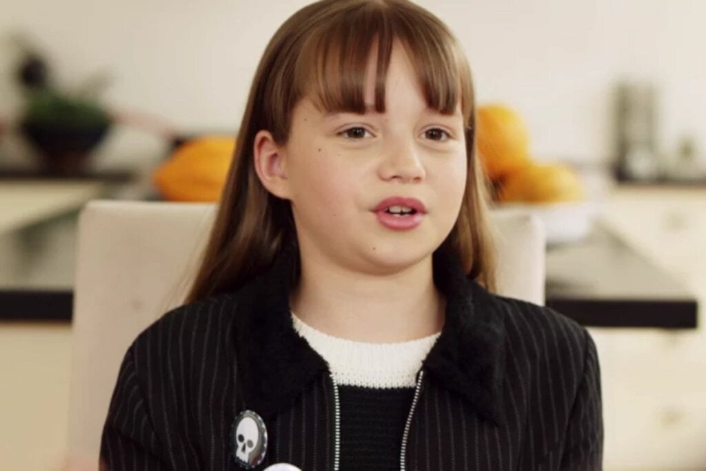 Menina com cabelo castanho com franja, usando roupa preta e sentada em um sofá em uma cena do filme 'O Baile das Bruxas'