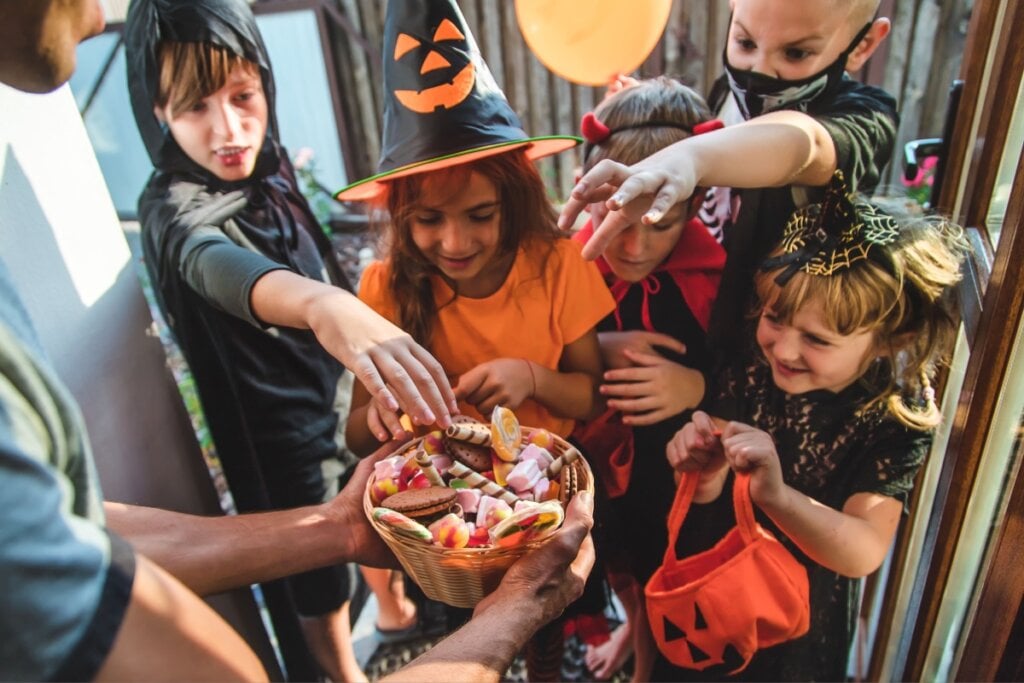 Crianças fantasiadas participando de "trick or treat", recebendo doces em um cesto durante a celebração de Halloween