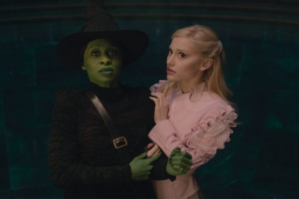 Elphaba e Glinda, protagonistas de "Wicked: Parte 1", lado a lado; uma é caracterizada com pele verde e chapéu pontudo, e a outra em trajes claros e delicados