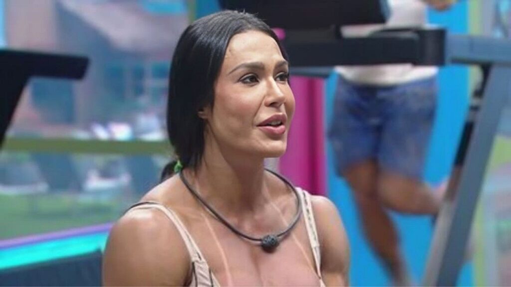 Gracyanne Barbosa está em um ambiente interno do reality show, possivelmente na academia, com um microfone preso ao pescoço. Ao fundo, é possível ver equipamentos de treino e parte de outro participante