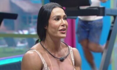 Gracyanne Barbosa está em um ambiente interno do reality show, possivelmente na academia, com um microfone preso ao pescoço. Ao fundo, é possível ver equipamentos de treino e parte de outro participante