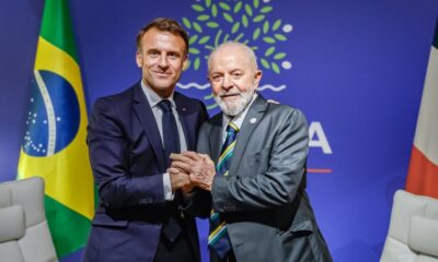 Lula e Macron de mãos dadas. Ao fundo, a bandeira do Brasil