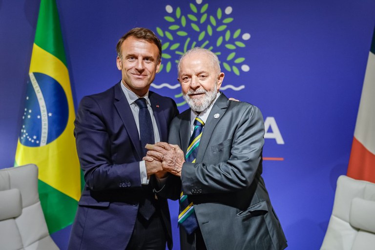 Lula e Macron de mãos dadas. Ao fundo, a bandeira do Brasil