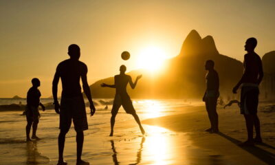 Grupo de pessoas jogando altinha na praia de Ipanema durante o pôr do sol, com o mar ao fundo e o Morro Dois Irmãos em destaque no horizonte. A luz dourada do sol cria uma atmosfera calorosa e vibrante