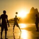 Grupo de pessoas jogando altinha na praia de Ipanema durante o pôr do sol, com o mar ao fundo e o Morro Dois Irmãos em destaque no horizonte. A luz dourada do sol cria uma atmosfera calorosa e vibrante