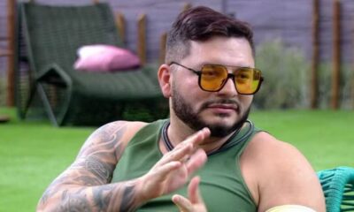 Marcelo Prata, participante do BBB 25 conversa na casa com óculos escuros bege e gramado verde ao fundo