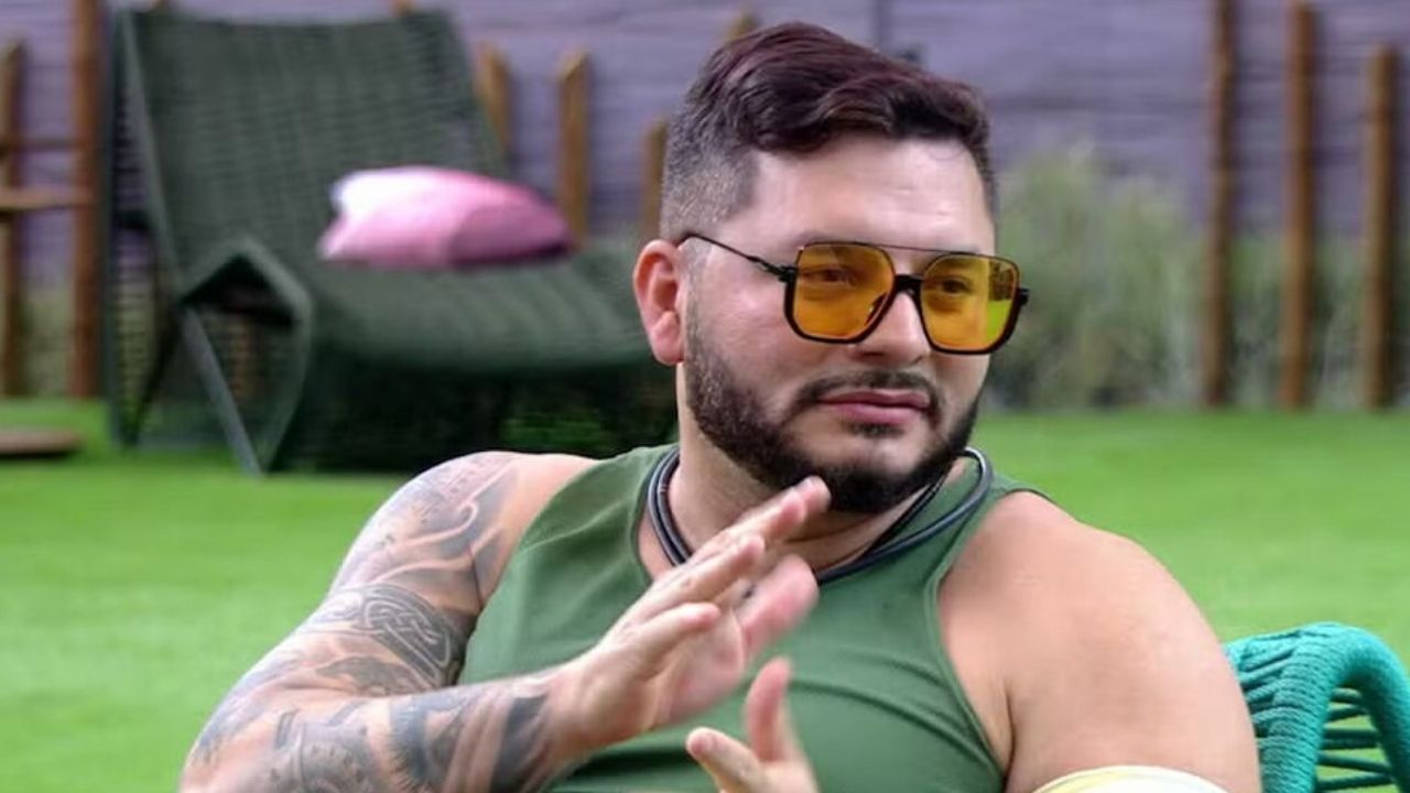 Marcelo Prata, participante do BBB 25 conversa na casa com óculos escuros bege e gramado verde ao fundo