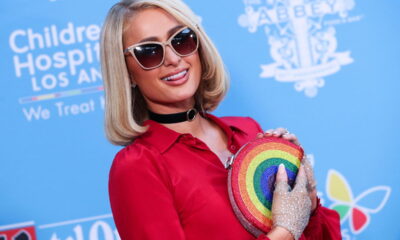 Paris Hilton de óculos escuros, segurando bolsa colorida e com fundo azul